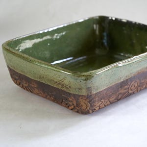 May include: A rectangular ceramic baking dish featuring a glossy sage green glaze on the interior and upper exterior, contrasted by a dark brown, unglazed lower exterior section decorated with a repeating embossed floral pattern around the sides.