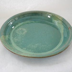 May include: A handmade ceramic pie plate with a turquoise glaze. The plate has a slightly irregular shape and a raised rim with a brown edge. The interior of the plate is a lighter shade of turquoise.