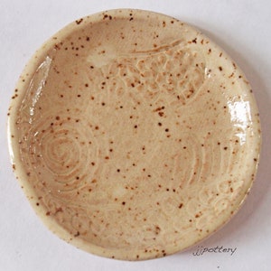 Puede incluir: Un plato pequeño, redondo, de cerámica con un esmalte moteado. El plato tiene un diseño en relieve de flores y espirales.