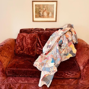 May include: A burgundy velvet couch with a patchwork quilt draped over it. The quilt is made of various fabrics in shades of pink, blue, and white.