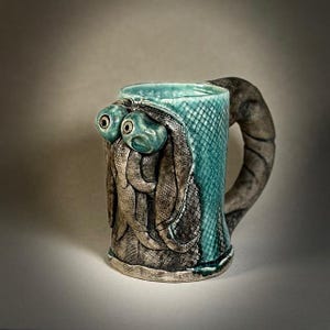 May include: A ceramic mug with a teal exterior and a textured, snake-like handle. The mug features a sculpted design of two creatures with large, round eyes. The mug's design includes a teal, scale-like pattern.