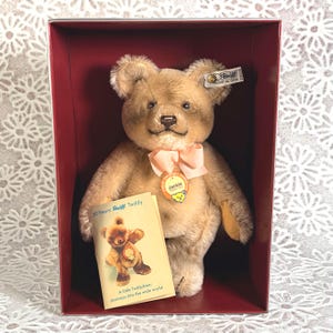May include: A light brown Steiff teddy bear with a peach-colored ribbon around its neck, a round yellow tag labeled Jackie, and a metal button in its ear. It is displayed in a dark red box with a small booklet titled 50 Years Steiff Teddy.