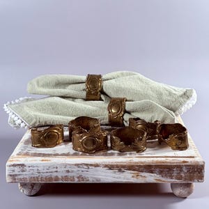 May include: A set of antique brass napkin rings with an ornate design, paired with sage green linen napkins. The napkins are folded and displayed on a distressed white wooden tray with rounded feet. The napkin rings have a vintage aesthetic.