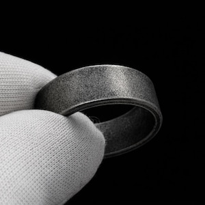 Könnte beinhalten: Nahaufnahme eines strukturierten, grauen Rings, der von einer behandschuhten Hand vor schwarzem Hintergrund gehalten wird. Der Ring hat ein flaches, breites Band mit leicht rauer Oberfläche. Die Farbe des Rings ist ein gedämpftes Grau.