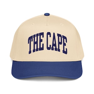 May include: A baseball cap with a cream-colored crown and a navy blue brim. The text "THE CAPE" is embroidered in navy blue across the front. The cap has a curved brim and a navy blue button on top.