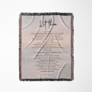 以下が含まれることがあります： フリンジ付きの縁取りが施された、クリーム色のスローブランケット。「Let Them」の文字がスタイリッシュなフォントで表示され、詩が添えられています。詩のテキストは、暗くエレガントなフォントです。