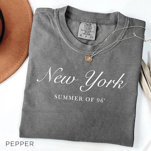 May include: A gray Comfort Colors t-shirt with the words "New York SUMMER OF 96'" in white script. A silver necklace with a round pendant rests on the shirt. A brown hat is partially visible.