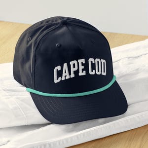 Puede incluir: Gorra de béisbol azul marino con el texto "CAPE COD" en letras blancas. La gorra tiene un detalle de cuerda verde turquesa a lo largo del borde. El sombrero está sobre una superficie blanca.