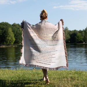 以下が含まれることがあります： フリンジ付きの縁取りが施された、プリントテキストが特徴の明るい色のブランケット。ブランケットは持ち上げられ、テキストが見えます。背景には湖、木、青い空が含まれています。ブランケットのテキストは「Let them love you.」と書かれています。