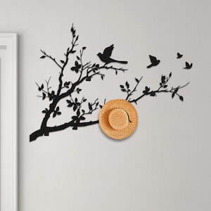 Könnte beinhalten: Schwarze Metall-Wandkunst mit einem Ast mit Vögeln und Blättern. Ein Strohhut hängt am Ast. Die Kunst ist an einer weißen Wand über einem Holzboden angebracht.