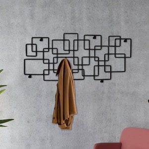 Könnte beinhalten: Ein schwarzer Metall-Kleiderständer mit geometrischem Design. Der Kleiderständer ist an einer grauen Wand montiert und hat eine braune Jacke daran hängen.