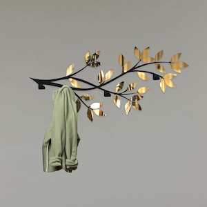 Può includere: Un appendiabiti da parete in metallo nero a forma di ramo d'albero con dettagli in foglia d'oro. Un maglione verde è appeso a uno degli ganci.
