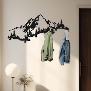 Op de afbeelding: Zwarte metalen wandhaak in de vorm van een bergketen met twee jassen die eraan hangen. De haak is gemonteerd op een witte muur met een houten deur en een houten tafel met een lamp en een kruk op de voorgrond.