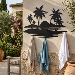 May include: A black metal wall-mounted hook rack featuring a silhouette of palm trees, a sun, and a coastal landscape. Three towels in beige, blue, and white are hanging from the hooks, which are integrated into the bottom of the metal design.