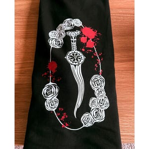 Può includere: T-shirt nero con grafica bianca di un pugnale circondato da rose e macchie rosse. Il disegno è centrato sul davanti della maglietta. La maglietta è piegata su una superficie di legno.