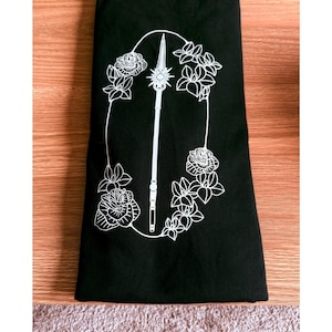 May include: Black t-shirt with a white floral and spear design. The design features a spear in the centre, surrounded by a floral wreath. The shirt is folded neatly on a wooden surface.