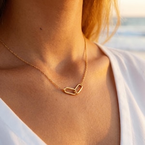 Puede incluir: Un delicado collar dorado con dos colgantes ovalados entrelazados. La cadena y los colgantes tienen un acabado pulido, que refleja la luz. El collar se lleva sobre una parte superior blanca, con un fondo suave y borroso.