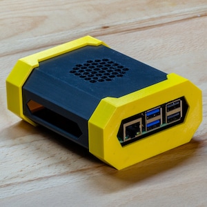 Könnte beinhalten: Ein schwarzes und gelbes 3D-gedrucktes Gehäuse für einen kleinen Computer mit einem Kühlventilator und mehreren Anschlüssen auf der Rückseite.