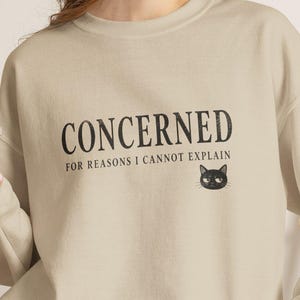 May include: A beige crewneck sweatshirt featuring the black text CONCERNED FOR REASONS I CANNOT EXPLAIN and a small illustration of a black cat face. The sweatshirt is paired with gray denim shorts, and a clear plastic cup with a straw is held.