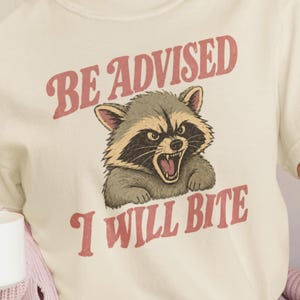 Könnte beinhalten: Ein cremefarbenes Kurzarm-T-Shirt mit einer Grafik eines knurrenden Waschbären und dem Text "BE ADVISED I WILL BITE", der in einer verwaschenen roten Schrift über und unter der Abbildung des Tieres gedruckt ist.