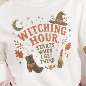 May include: A white short-sleeved t-shirt featuring a graphic print with the text "WITCHING HOUR STARTS WHEN I GET THERE". The design includes a witch hat, a crescent moon, a boot, a lipstick tube, and autumn leaves in orange and brown tones.