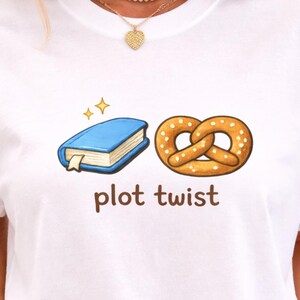 May include: White t-shirt featuring a cartoon illustration of a blue book with gold stars and a pretzel. The words "plot twist" are written in brown font below the images. The shirt is a casual style.
