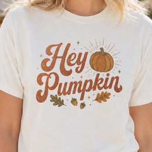 May include: A cream-colored short-sleeve t-shirt featuring the text Hey Pumpkin in a brown retro font, accompanied by a graphic of a pumpkin, small stars, an acorn, and two oak leaves centered on the front of the garment.