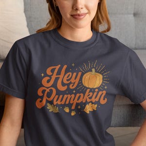 May include: A dark gray short-sleeved t-shirt featuring the orange text "Hey Pumpkin" in a retro font. The design includes a central orange pumpkin illustration with radiating lines, small stars, two acorns, and two oak leaves printed on the front.