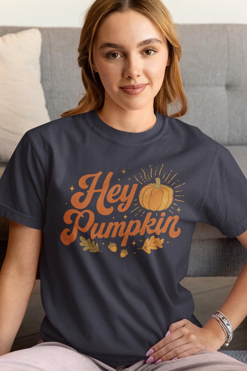 May include: A dark gray short-sleeved t-shirt featuring the orange text "Hey Pumpkin" in a retro font. The design includes a central orange pumpkin illustration with radiating lines, small stars, two acorns, and two oak leaves printed on the front.