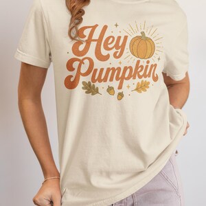 May include: A cream-colored short-sleeve t-shirt featuring the text Hey Pumpkin in a brown retro font. The graphic includes a central orange pumpkin illustration with radiating sunburst lines, surrounded by small brown acorns and oak leaves.