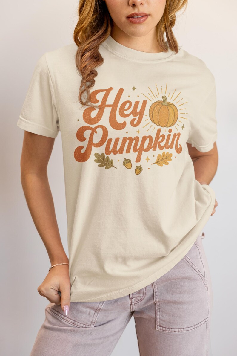 May include: A cream-colored short-sleeve t-shirt featuring the text Hey Pumpkin in a brown retro font. The graphic includes a central orange pumpkin illustration with radiating sunburst lines, surrounded by small brown acorns and oak leaves.