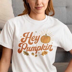 May include: A white short-sleeved t-shirt featuring a graphic print with the text "Hey Pumpkin" in a brown retro font. The design includes an orange pumpkin illustration with radiating lines, small stars, two brown acorns, and two oak leaves.
