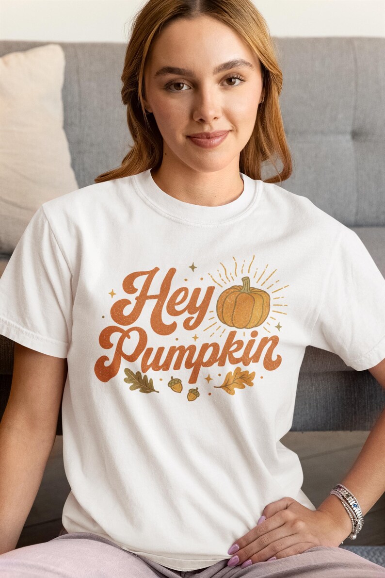 May include: A white short-sleeved t-shirt featuring a graphic print with the text "Hey Pumpkin" in a brown retro font. The design includes an orange pumpkin illustration with radiating lines, small stars, two brown acorns, and two oak leaves.
