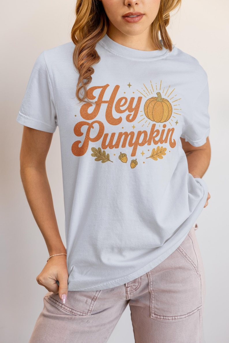 May include: A light blue short-sleeve t-shirt featuring the orange text "Hey Pumpkin" in a retro font. The graphic includes an orange pumpkin illustration with radiating sunburst lines, small stars, and several brown oak leaves and acorns.