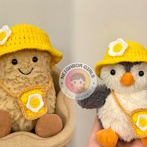 May include: Two plush toys wearing yellow crocheted hats with white flower accents. The toys are a peanut-shaped character and a penguin. Both toys have a yellow crocheted strap with a white flower accent around their necks.