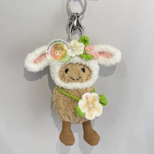 Könnte beinhalten: Ein Schlüsselanhänger mit einem Schaf aus Plüsch, das ein weiß-cremefarbenes Lammkostüm trägt. Das Schaf trägt einen Blumenkranz und hält eine Blume. Der Schlüsselanhänger hat einen silbernen Verschluss und einen Ring. Der Text "NEIGHBOR GIRLS" steht auf dem Kopf des Schafes.