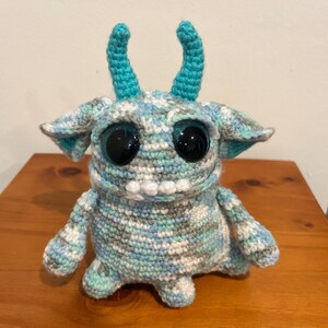 May include: A handmade crochet monster doll with large, black, glittery eyes and a toothy grin. The doll features blue, gray, and white yarn, with turquoise horns and ear accents. The doll is sitting on a wooden surface.