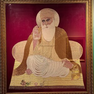Puede incluir: Una obra de arte con cuentas enmarcada que representa a una figura barbuda con turbante y halo, sentada sobre una estera amarilla sobre un fondo rojo intenso. La figura viste una túnica clara y un chal marrón, con una mano alzada en bendición.