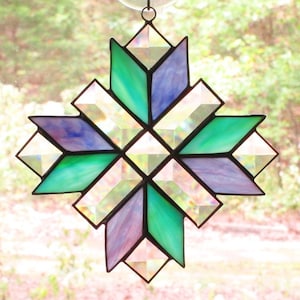 May include: A stained glass suncatcher with a geometric design in shades of green, purple, and clear. The suncatcher is shaped like a star and has a hanging loop at the top.