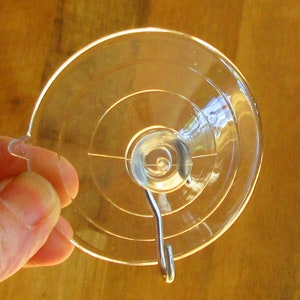 May include: A clear plastic suction cup with a metal hook. The suction cup has a circular shape with concentric circles.