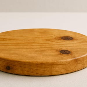 May include: A round, wooden cutting board with a light brown finish. The board has a smooth surface with two dark knots and a rounded edge. The wood grain is visible, and the board appears to be new.