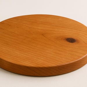 May include: A round, wooden cutting board with a warm, reddish-brown hue. The board has a smooth surface with visible wood grain and a knot detail. The edges are rounded, and the board appears to be about 1 inch thick.