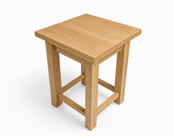 May include: A square, light-colored wooden stool with a natural finish. The stool has a square top and four sturdy legs, with a cross-brace design near the base for added support. The wood grain is visible.