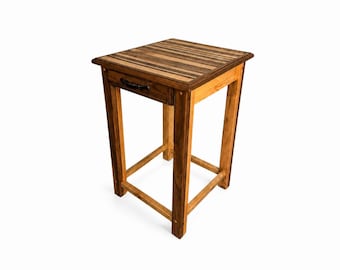 May include: A small wooden side table with a drawer. The tabletop is made of alternating light and dark wood strips. The drawer has a black handle. The legs are square and the table is a warm brown color.