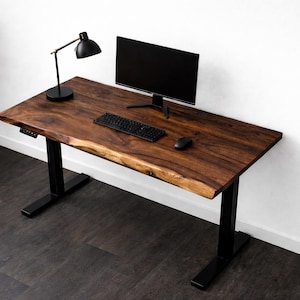 May include: A dark brown wooden desk with a live edge, supported by black metal legs. A black computer monitor, keyboard, and mouse sit on the desk. A black desk lamp is on the left. The desk is in front of a white wall.