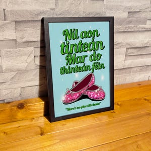 K&ouml;nnte beinhalten: Ein gerahmter Druck mit blauem Hintergrund und gr&uuml;nem Text in Irisch-G&auml;lisch, der "Nil aon tintean mar do thintean fein." lautet. Der Text ist von zwei pinkfarbenen, glitzernden Schuhen umgeben. Der Text unter den Schuhen lautet "Es gibt keinen Ort wie zu Hause."