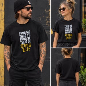 K&ouml;nnte beinhalten: Ein schwarzes T-Shirt mit kurzen &Auml;rmeln und wei&szlig;em Text, der THUG M&Eacute;, THUG TU, THUG SE, THUG SI lautet, wobei die Worte THUG LIFE darunter in einer gelben Schrift im gotischen Stil gedruckt sind. Die R&uuml;ckseite des T-Shirts ist schlicht schwarz.