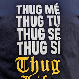 K&ouml;nnte beinhalten: Ein schwarzes T-Shirt mit wei&szlig;em Text, der THUG ME, THUG TU, THUG SE, THUG SI in einer gestapelten serifenlosen Schrift anzeigt, gefolgt von den Worten THUG LIFE, die in einer fetten, gelben, gotischen Schrift unter dem Text stehen.
