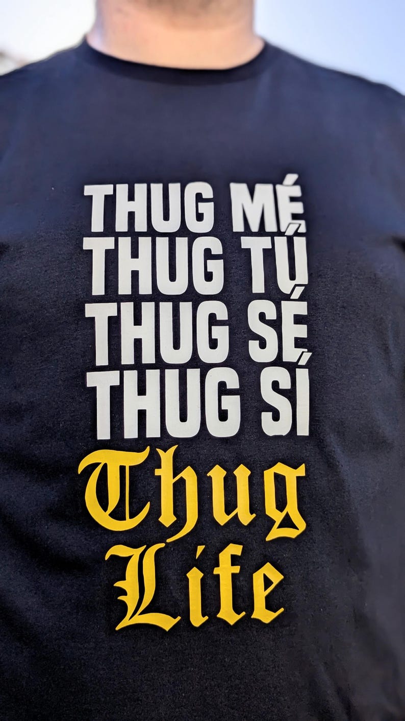 K&ouml;nnte beinhalten: Ein schwarzes T-Shirt mit wei&szlig;em Text, der THUG ME, THUG TU, THUG SE, THUG SI in einer gestapelten serifenlosen Schrift anzeigt, gefolgt von den Worten THUG LIFE, die in einer fetten, gelben, gotischen Schrift unter dem Text stehen.