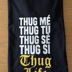 K&ouml;nnte beinhalten: Ein schwarzes T-Shirt mit kurzen &Auml;rmeln und wei&szlig;em Text, der THUG ME, THUG TU, THUG SE, THUG SI lautet, gefolgt von dem gelben Text THUG LIFE in einer gotischen Schriftart. Der Innenkragen zeigt das wei&szlig;e Markenlogo Tucutkun auf schwarzem Grund.
